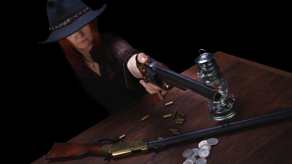 wild west girl shooting from revolver gun at the table with ammunition and silver coins on black background