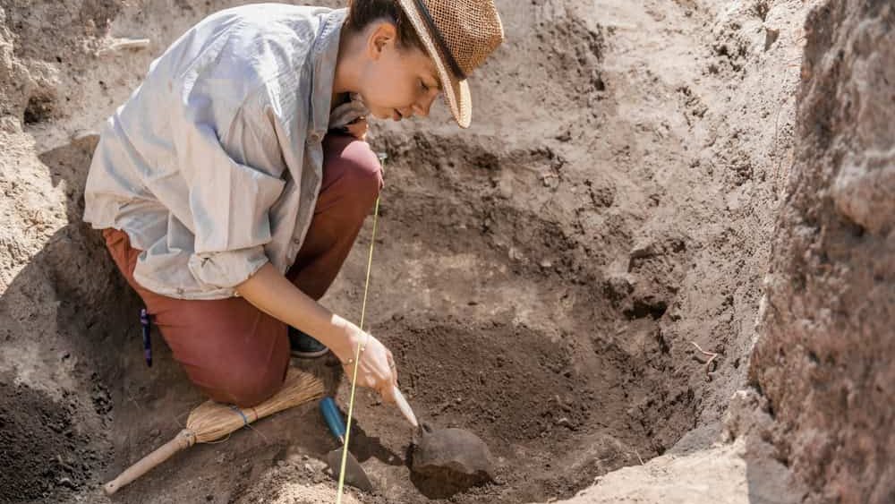 Archaeologist digging with hand trowel, recovering ancient pottery object from an archaeological site.