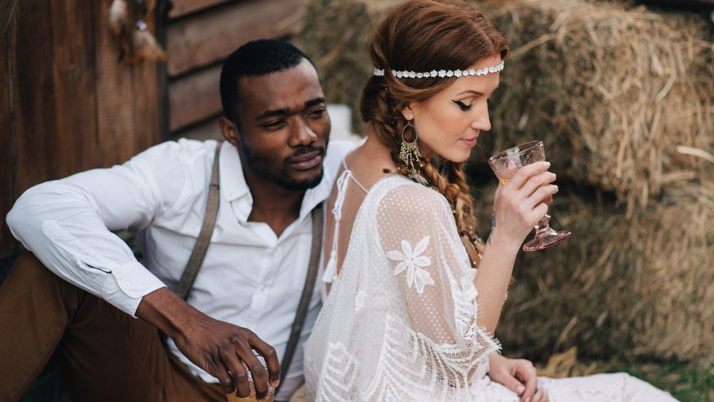Boho style wedding. African-American bridegroom and Caucasian bride drink wine from glasses on a picnic.