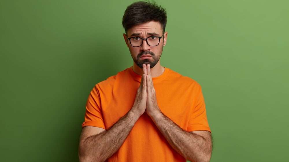 Sad European man with hopeless upset expression prays and has hope for better, guilty look, asks for forgiveness, feels really sorry, presses hands together says please forgive me wears orange t shirt