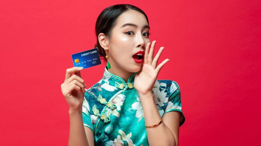 Cute Asian woman wearing oriental qipao dress showing credit card cupping hand and saying on red studio background for Chinese new year cashless shopping concepts