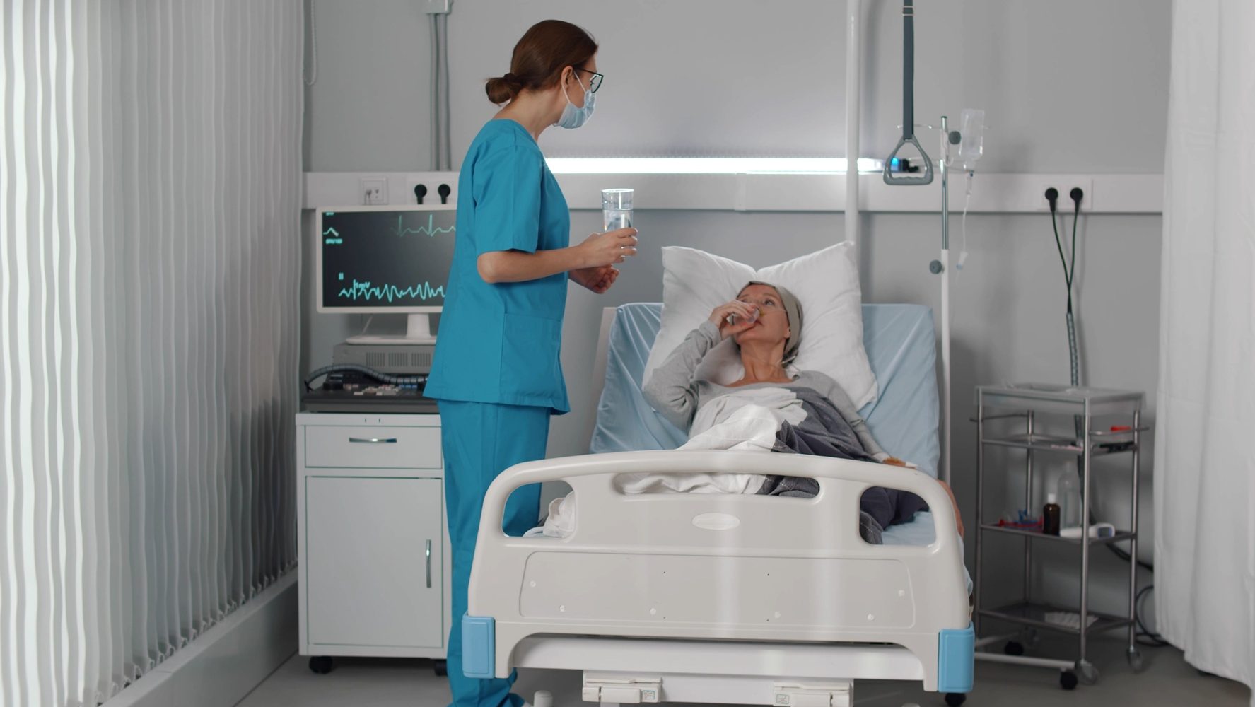 Nurse giving medication and water to cancer patient lying in bed at hospital. Medical worker in scrubs and safety mask giving pills to sick woman in hospital ward