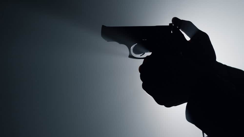 Silhouette of man hand holding gun indoors. Closeup unknown criminal arm cocking trigger in spotlight background. Unrecognizable male person shooting firearm in dark.