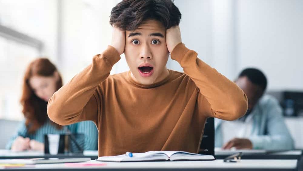 Portrait,Of,Shocked,Asian,Male,Student,Sitting,At,Desk,In