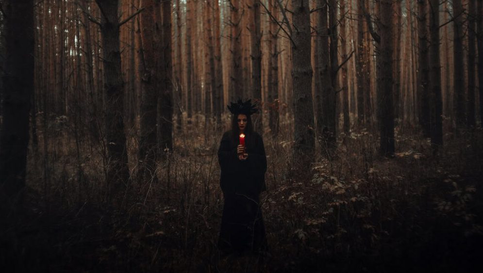 witch in a black costume holds candles in a dark forest