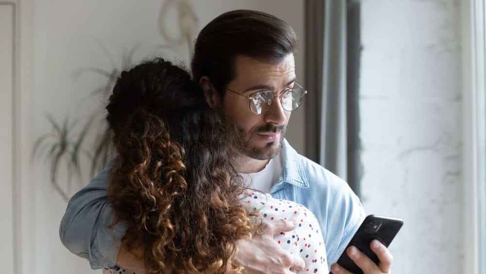 Young husband hugs wife holds cellphone check messages in social media suspect her in treason. Broken trust in relationships, jealous boyfriend, mistrust in relations, crisis of family life concept