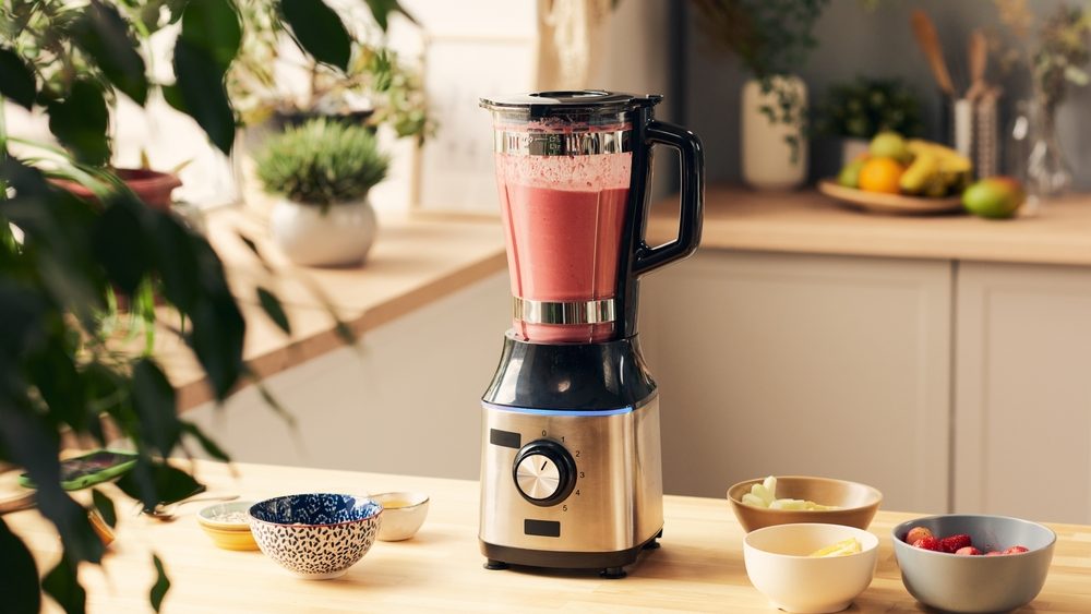 Electric blender with fresh homemade fruit smoothie and group of bowls containing ingredients on wooden kitchen table