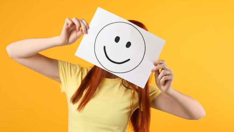Woman hiding behind sheet of paper with happy face on yellow background