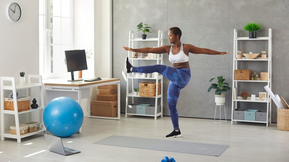 Happy black woman having sports workout at home. Active young lady standing in living room, watching online fitness workout lesson on modern laptop computer and doing leg swings exercise
