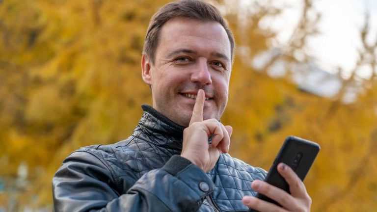 Portrait of mysterious attractive guy using gadget showing shh sign in park outdoors. Millennial man holding mobile phone doing silence gesture