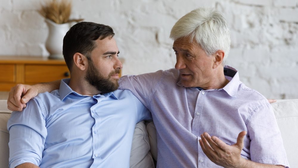 Elder grey haired loving father giving support and advice to grownup son. Senior parent and adult child talking on couch at home, discussing family life, keeping trusting relationships