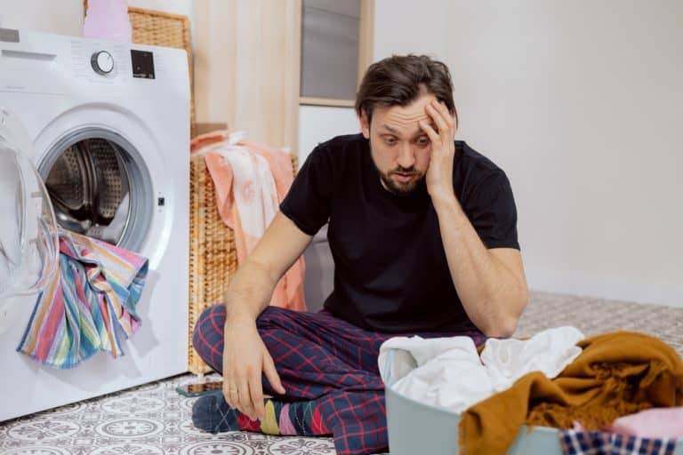 Guy sits on the laundry room floor with the washing machine open, throwing all the clothes into the drum, guy doesn't know how to do housework, face crumpled, catches head with hand.