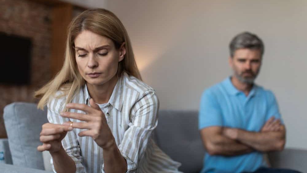 Offended sad adult caucasian woman takes off ring and ignores unhappy husband at conflict in living room interior. Problems in relationships, emotions after quarrel at home, divorce and breakup