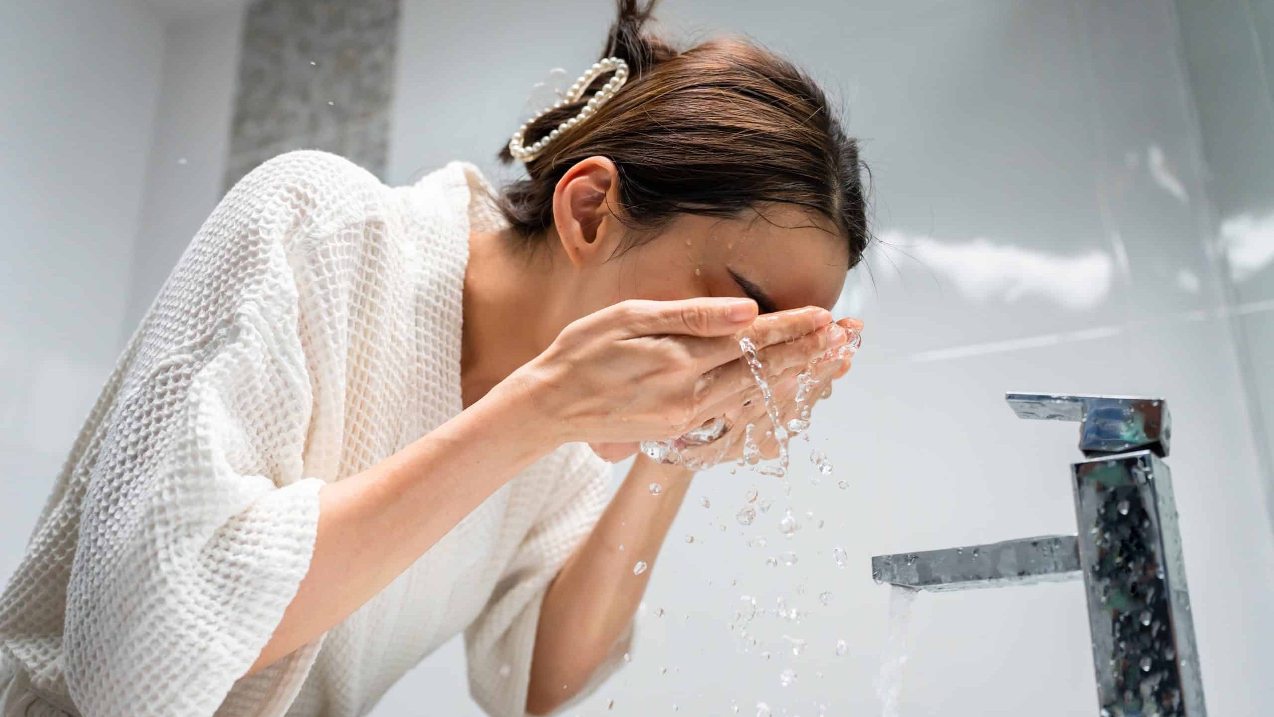 Asian beautiful woman washing her clean face with facial foam and water. Attractive female in bathrobe washing face for healthy beauty treatments and skin care then looking at the mirror in bathroom.