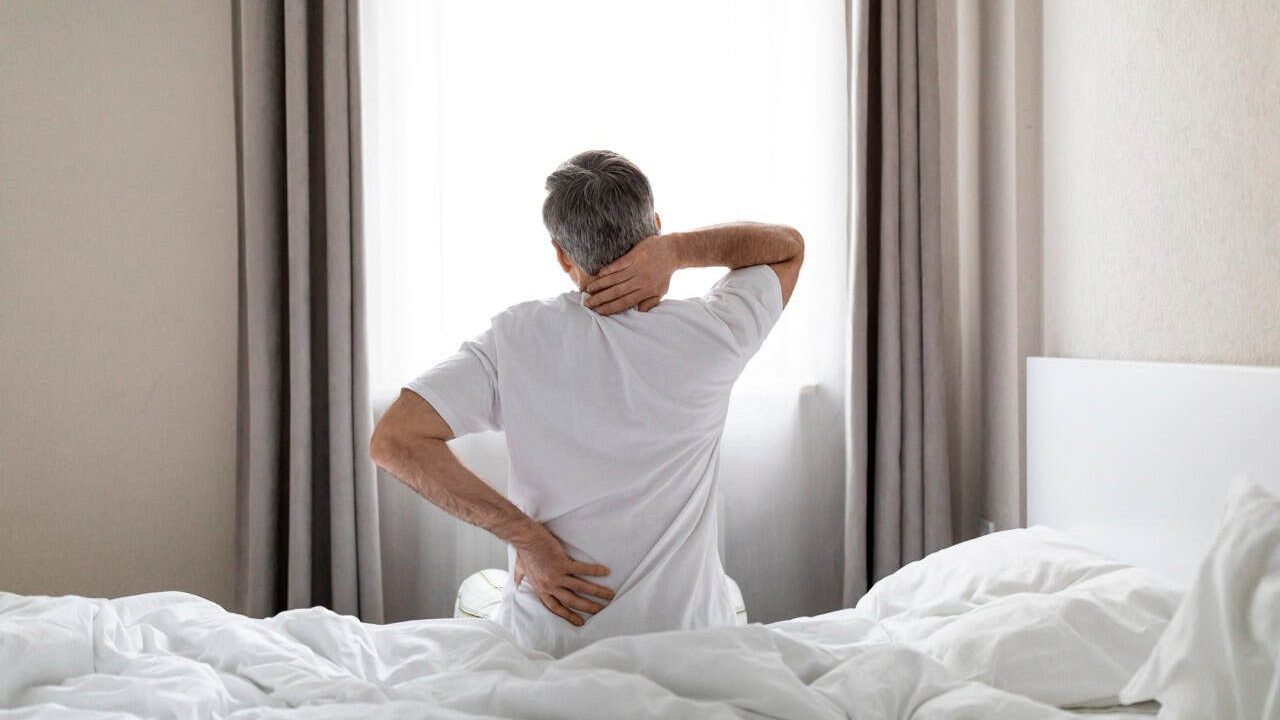 Unrecognizable mature grey-haired man wearing white pajamas sittting on bed at home in the morning, touching lower back and neck, suffering from muscle strain after waking up, copy space