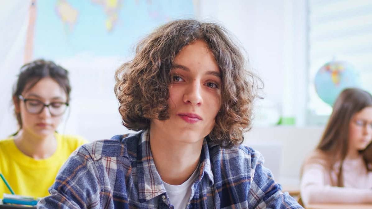 Portrait shot of Caucasian teenage guy with long hair looking at camera while sitting at lesson in classroom. Close up of face of teen boy. School life concept. Learning at class.