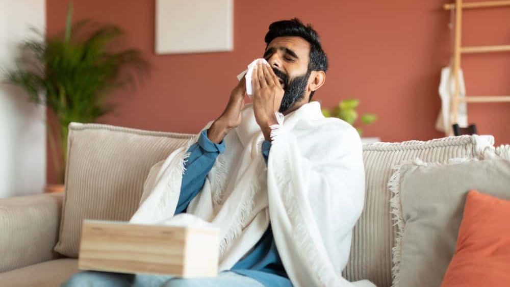 Sick Indian Man Blowing Runny Nose In Paper Tissue Sitting On Sofa At Home, Suffering From Rhinitis And Sinusitis Symptoms. Ill Middle Eastern Guy Sneezing Having Cold Posing Wrapped In Blanket