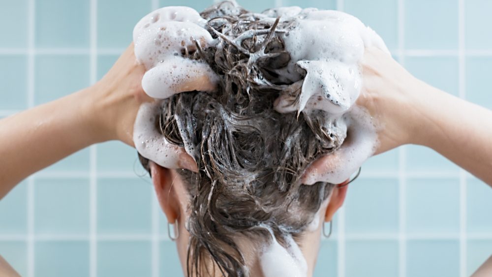 Girl washes her hair with shampoo on blue background, back view