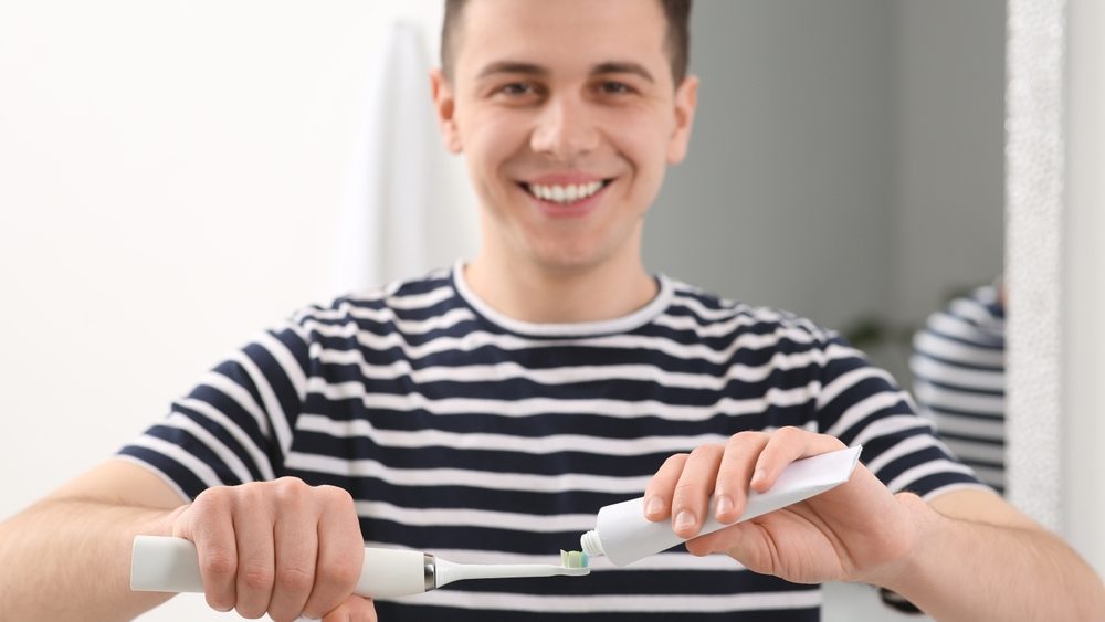 Man squeezing toothpaste from tube onto electric toothbrush in bathroom, selective focus