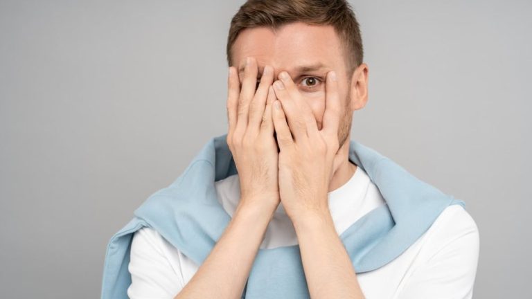 Shy modest playful guy hiding closing face with hands one eye looking at camera on isolated grey background. Portrait bashful timid coy man burns with shame feels awkward. Sincere emotions concept.