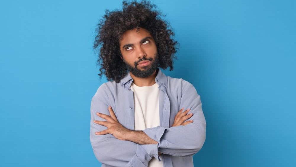 Young doubtful funny Arabian man rolls eyes and looks up with offense crossing arms in front of chest after insult or inappropriate remark from boss posing on isolated blue background.