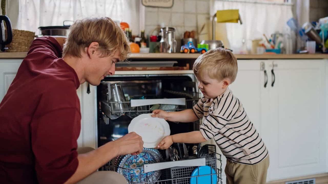 Little boy helping father to load dishwasher after breakfast. Cleaning the kitchen before leaving to work and daycare. Family morning routine.