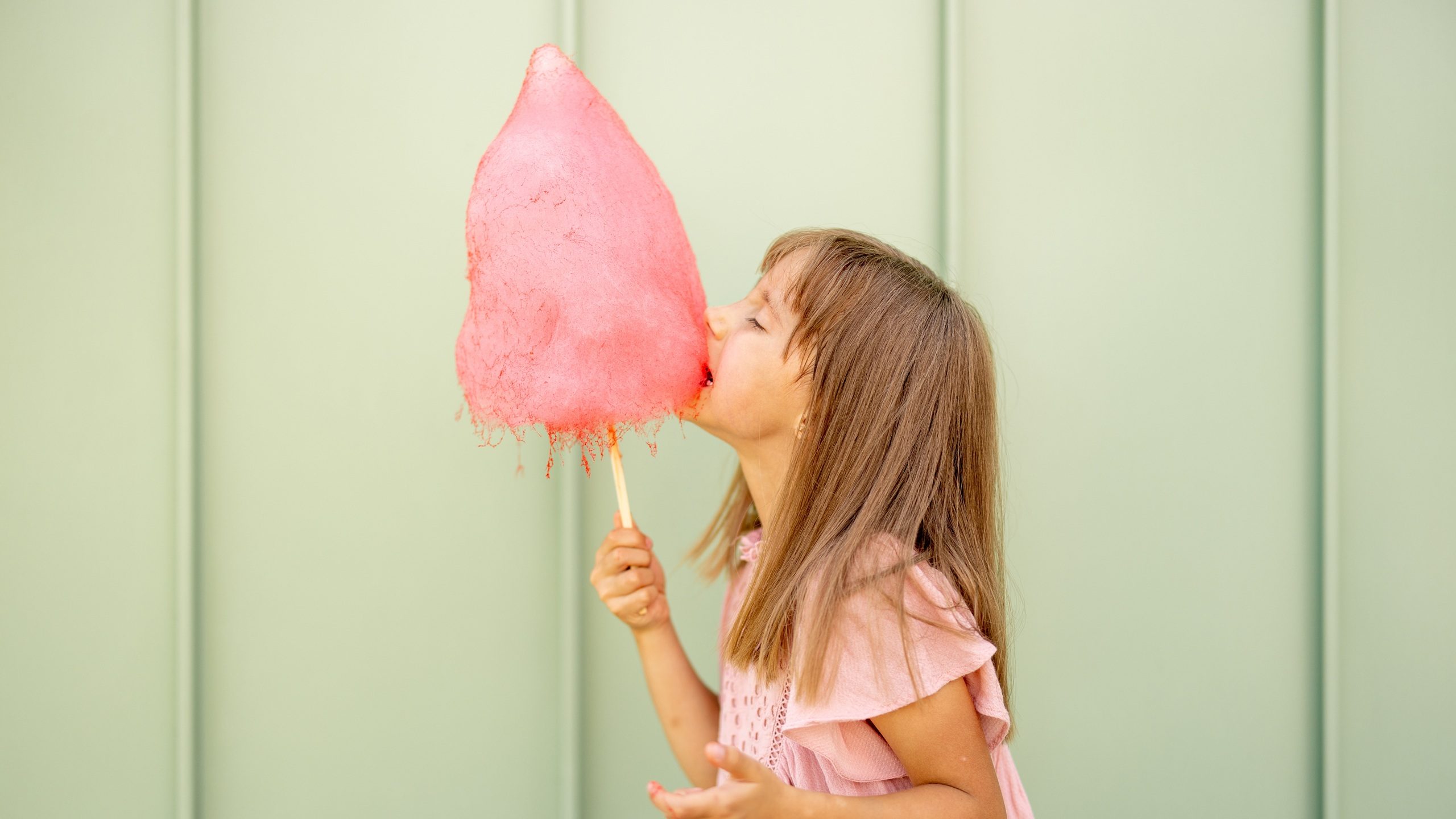 Portrait of a little girl with pink cotton candy on green wall background outdoors. Happy kids visiting amusement park and having fun