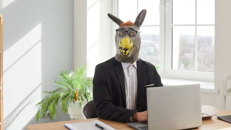 Businessman in donkey head mask using laptop computer in office. Funny stupid male office employee in suit, rubber mask and glasses sitting at desk with laptop and looking at camera