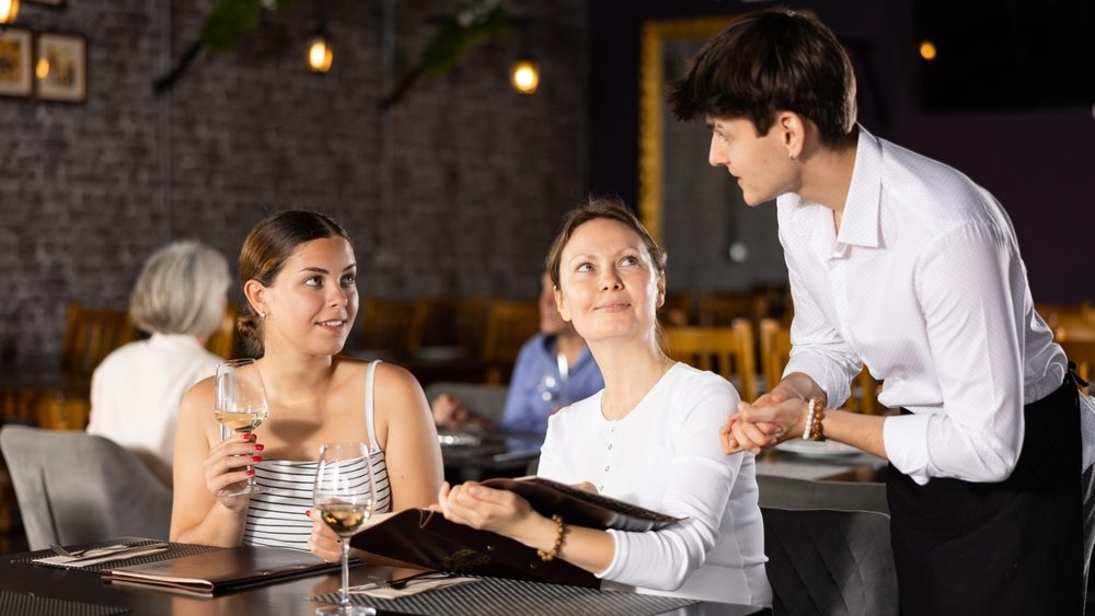 Young male waiter in uniform takes order from two women in restaurant