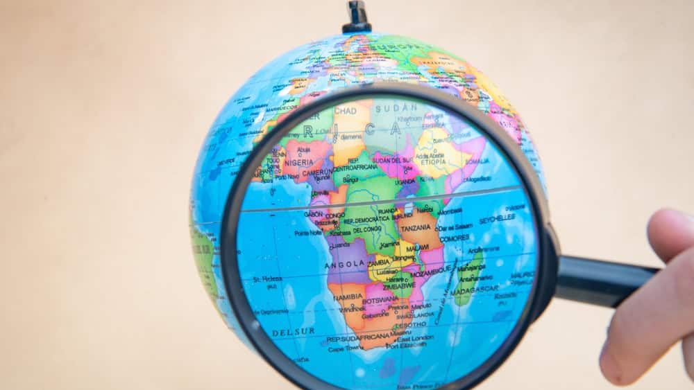 Photo of a globe with a magnifying glass magnifying countries, clear background with a hand placing the magnifying glass.
