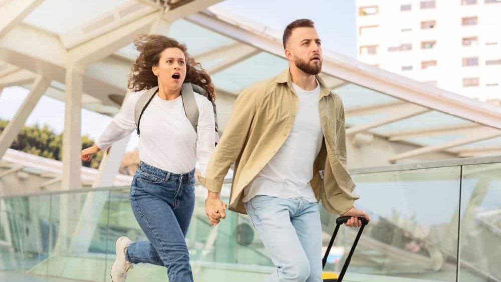 Stressed young couple running with luggage in airport terminal, late for their flight, in motion shot of emotional man and woman in rush, holding hands while going to flight gate, free space
