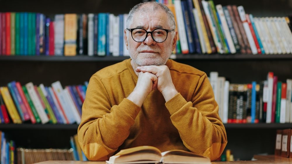 Amidst the book-lined aisles of the library, a dignified senior man, adorned with glasses and a beard, exudes an aura of intellectual curiosity and scholarly wisdom.