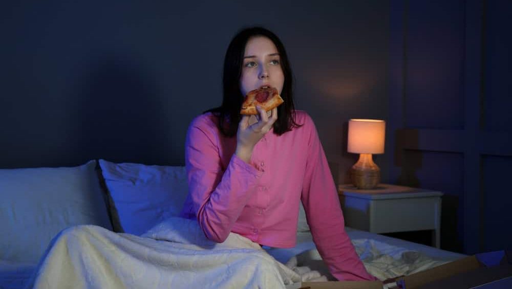 Young,Woman,Eating,Pizza,While,Watching,Tv,On,Bed,At