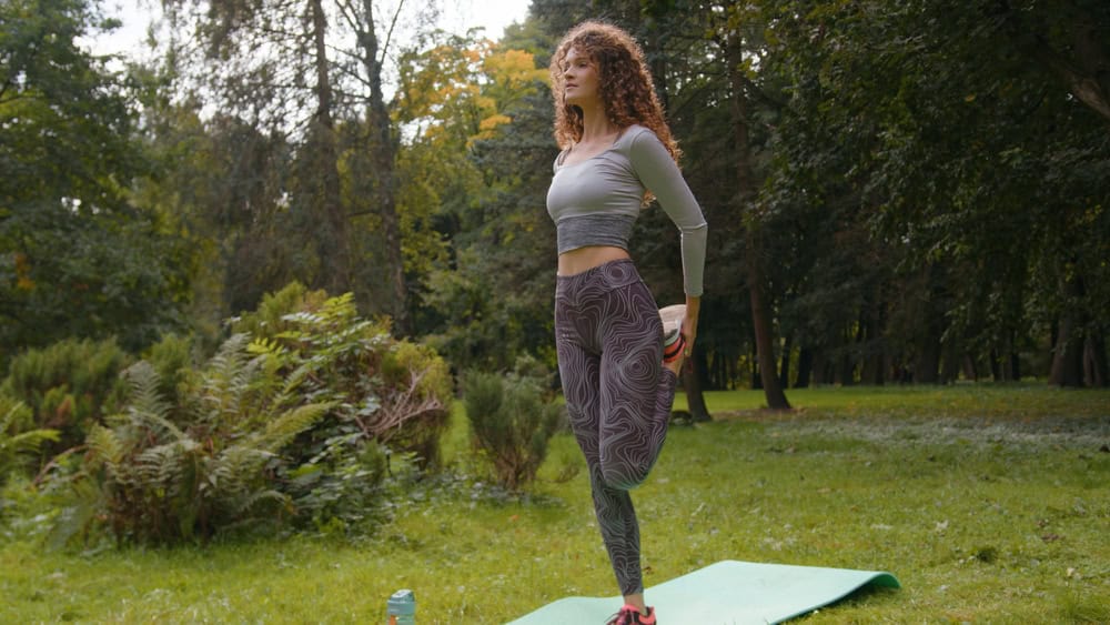 Caucasian sporty woman standing on one leg rotate hip warms up hips joints bend knee back sport stretching balance stretch workout yoga in park outdoors girl doing exercise fitness hold hands to sides