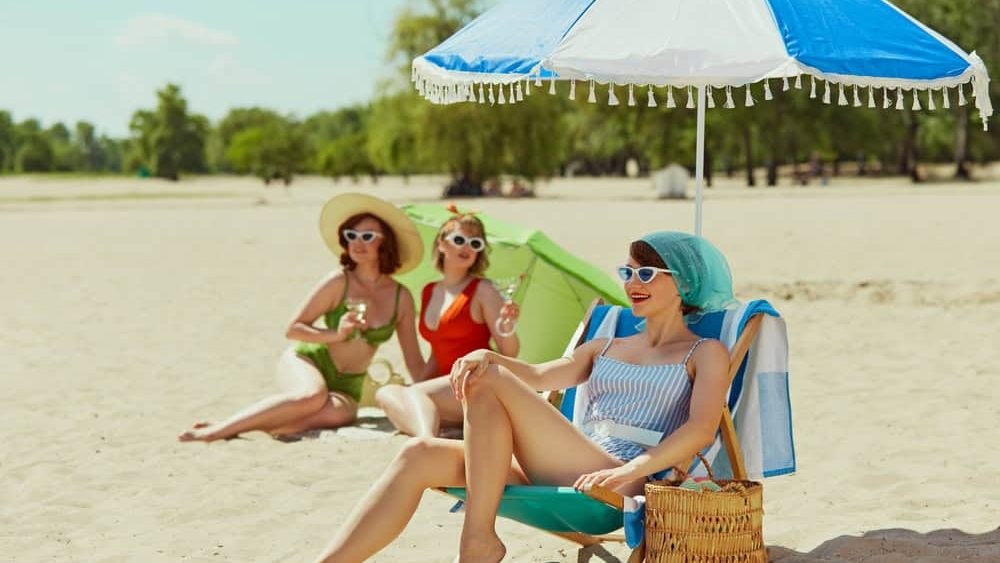 Three women in retro swimwear enjoying sunny beach day. One sits under umbrella with drink, while other two relax with green parasol. Vintage summer vibes. Concept of summer holiday, vacation, relax