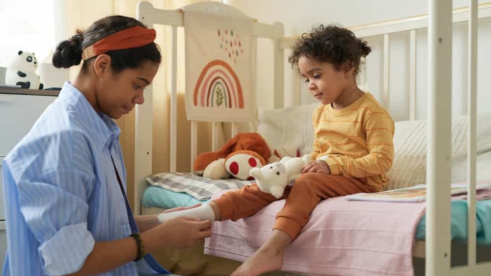 Side view portrait of caring older sister dressing little girl in cozy nursery and helping putting on socks