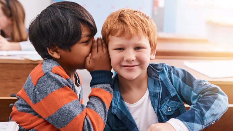 Education, naughty and secret with boy students in classroom together for learning or study. Gossip, question and whisper with children friends at desk in class for development, future or growth