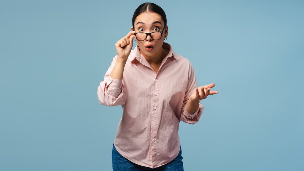 Young Asian woman is adjusting her glasses with a shocked expression on her face, gesturing with her hand as if she has just heard surprising news