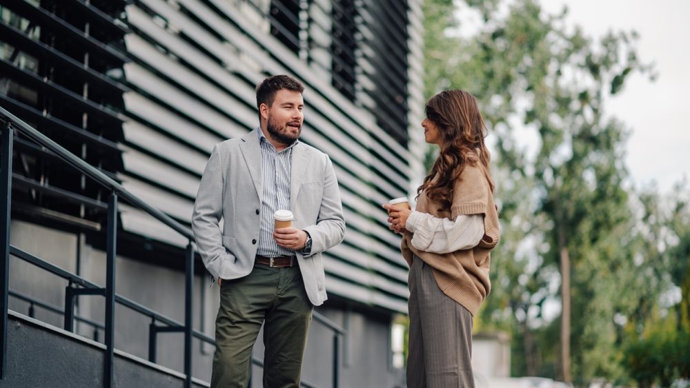Two business partners enjoying coffee outside a modern office building, taking a break from work and chatting happily in the city