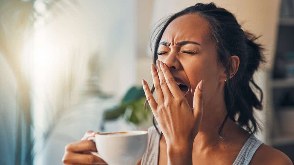 Tired woman, yawn and morning with coffee or waking up for lazy, sleepy or nap at home. Young, female person or awake with cup of caffeine for beverage, drink or cappuccino in daily routine at house