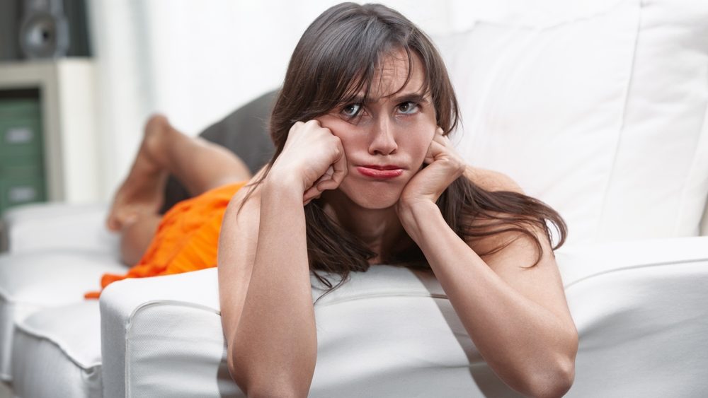 Tired woman lies on her cozy sofa at home, looking bored and sad. She seems lost in thought, feeling lonely in her casual apartment. This captures a moment of dull routine and discontent