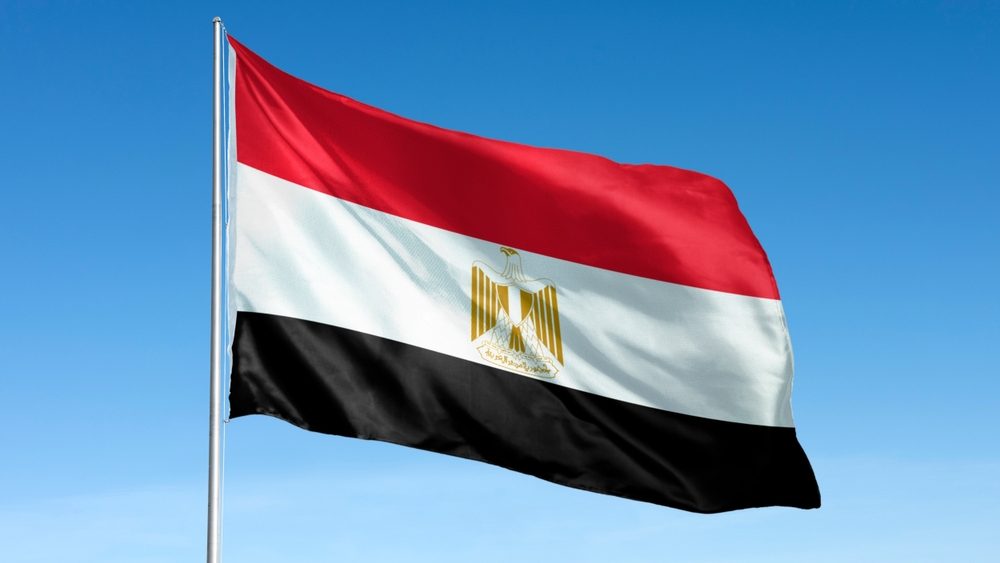 The image shows the Egyptian flag waving against a clear blue sky. The flag of Egypt features red, white, and black horizontal stripes with a golden eagle emblem. Flag of Egypt.