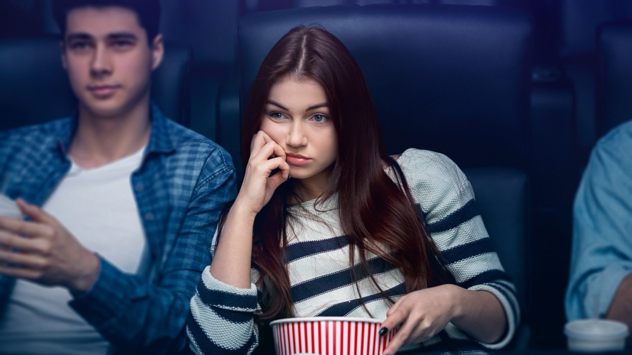 Boring date concept. Weary young girl with boyfriend watching movie in dark cinema