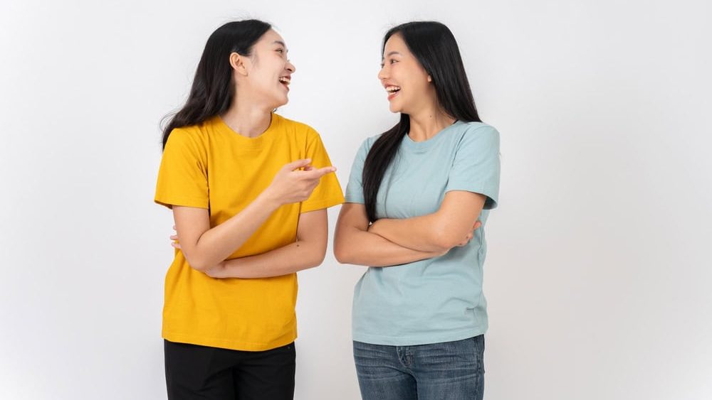 Two cheerful, lively Asian female friends in casual clothes standing against a white studio background, enjoying talking and laughing together. best friend, friendship, relationship