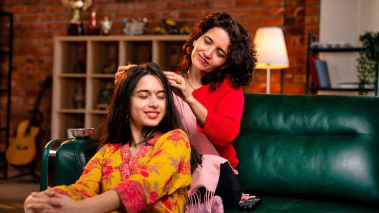 Indian Asian mother in her fifties lovingly giving an oil head massage to her pretty young daughter while sitting together on a sofa at home, expressing care and affection