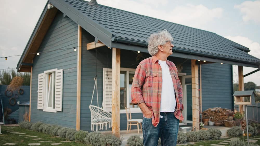 Older Caucasian man with grey hair and glasses looking at something in distance. Covering eyes with one hand from bright sunlight. One story building in background. Wearing checkered shirt.