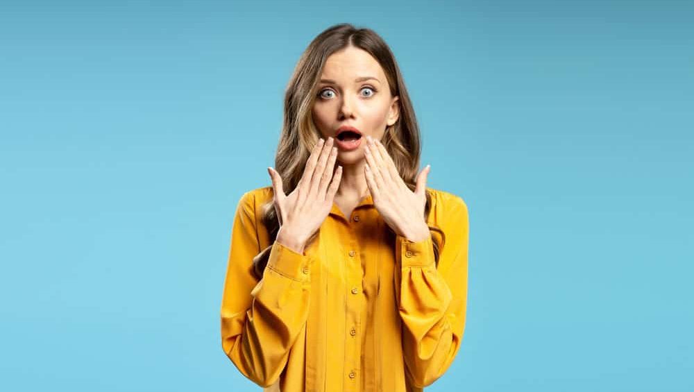 Studio portrait of surprised businesswoman covering her mouth with hands, expressing astonishment and disbelief on blue background