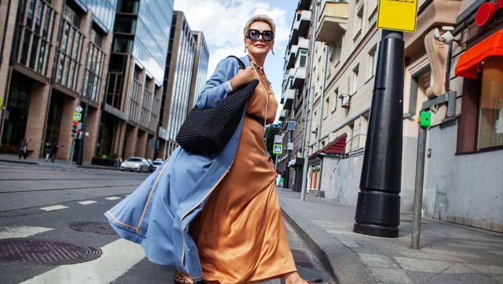 Stylish Mature Woman Walking City Street In Fashionable Clothes, Wearing Long Beige Silk Dress, Blue Cape Coat, Carrying Large Black Handbag, Sunglasses And Trendy Loafers