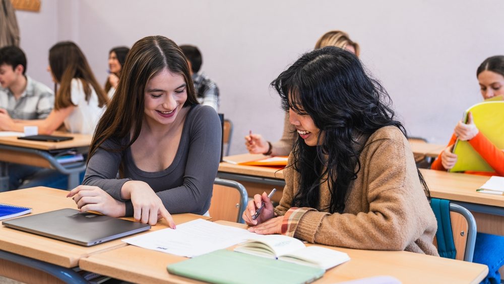 Two high school students are smiling and working together on a project in a classroom, using a laptop and taking notes