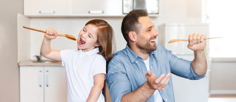 Father And Little Daughter Having Fun Cooking Together Singing Holding Spoons Like Microphones In Kitchen At Home. Funny Food Preparation